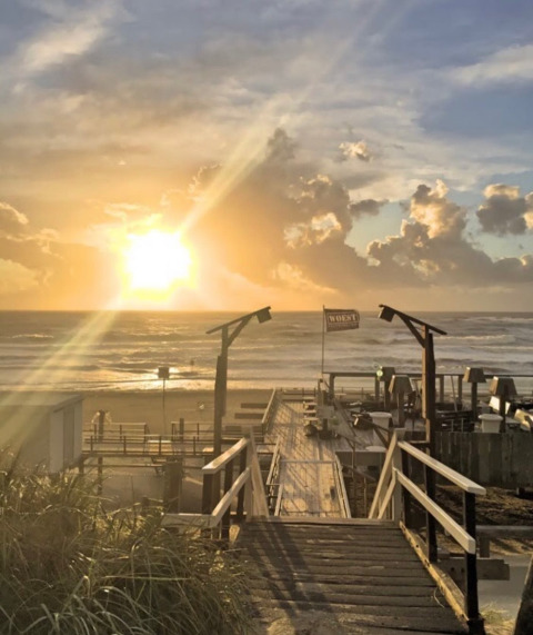 Amanecer en la playa desde un glamping con pasarela de madera, sombrillas y nubes de fondo.