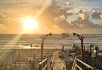 Amanecer en la playa desde un glamping con pasarela de madera, sombrillas y nubes de fondo.