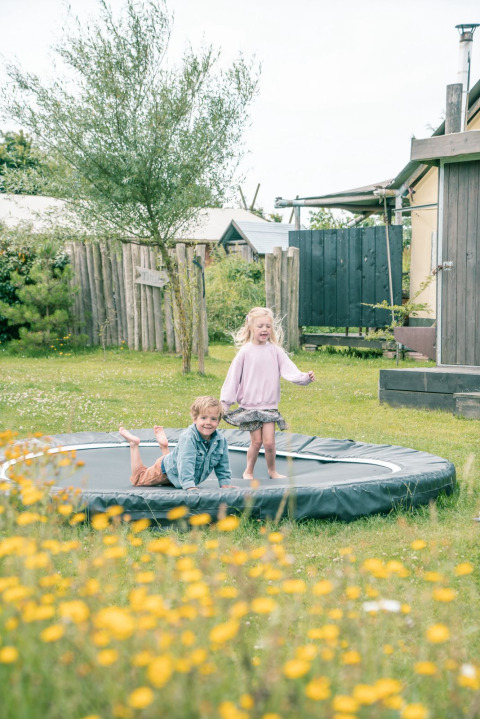Twee kinderen spelen op een ingegraven trampoline tussen veldbloemen bij een glampingverblijf.