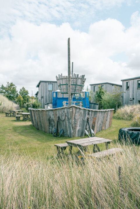 Photo d’un hébergement glamping avec une aire de jeux en bois en forme de bateau, entourée de cabanes et d’herbe.
