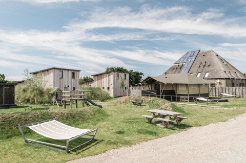 Buitenbeeld van een moderne lodge met houten cabines, picknickbanken, hangmatten en speeltuigen.