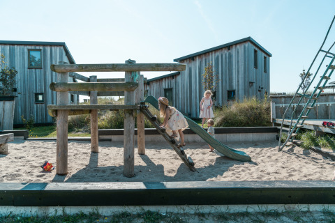 Bambini giocano su un parco giochi con sabbiera in un glamping circondato da casette di legno.