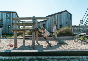 Des enfants jouent sur une aire de jeux avec bac à sable dans un hébergement glamping entouré de cabanes.
