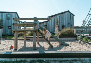 Kinder spielen auf einem Spielplatz mit Sandkasten bei einer Glamping-Unterkunft mit Holzhütten.