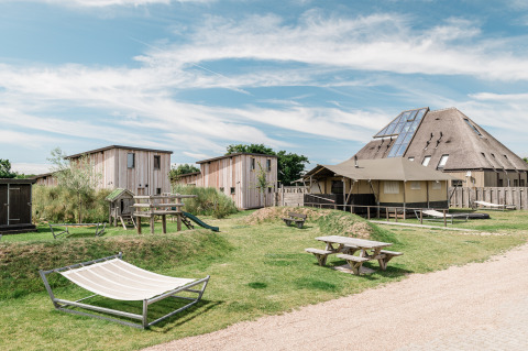 Alojamiento glamping con cabañas modernas de madera, área de juegos, mesas y hamaca en un día soleado.