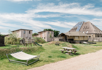 Alojamiento glamping con cabañas modernas de madera, área de juegos, mesas y hamaca en un día soleado.
