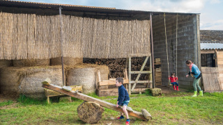 Niños juegan en un subibaja y columpio frente a un granero con pacas de heno en Feather Down L'Étable des Mauges en Anjou.