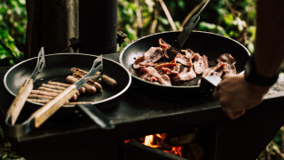 Persona cocina desayuno al aire libre, friendo tocino y salchichas en sartén sobre llama abierta en el bosque.