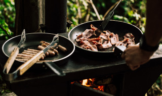 Persona cocina desayuno al aire libre, friendo tocino y salchichas en sartén sobre llama abierta en el bosque.