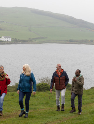 Fire personer går langs en grøn eng ved en sø nær Tunbridge Wells med bakker og huse i baggrunden.