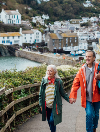 Pareja mayor paseando de la mano en un sendero junto al mar en un parque vacacional con glamping y pueblo.