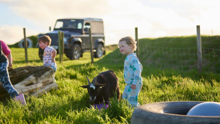 Niños en pijama juegan en el césped con una cabra en Feather Down Treganhoe Farm, suroeste de Inglaterra.
