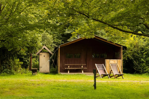 Et safari telt med to liggestole foran, et udendørs bord og stole, omgivet af grønne træer og natur.