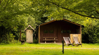 A safari tent with two deck chairs, an outdoor table, and lush green trees in a peaceful nature setting.