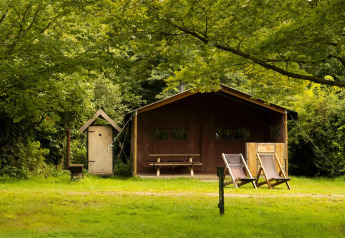Et safari telt med to liggestole foran, et udendørs bord og stole, omgivet af grønne træer og natur.