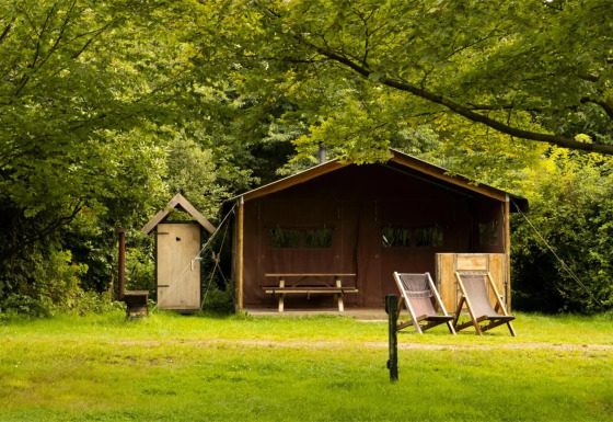 A safari tent with two deck chairs, an outdoor table, and lush green trees in a peaceful nature setting.