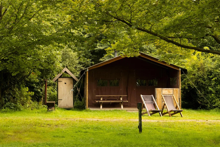 A safari tent with two deck chairs, an outdoor table, and lush green trees in a peaceful nature setting.