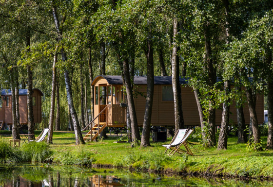 Gipsy-vogn med terrasse i en skov ved søen, omgivet af træer og liggestole i grønne omgivelser.