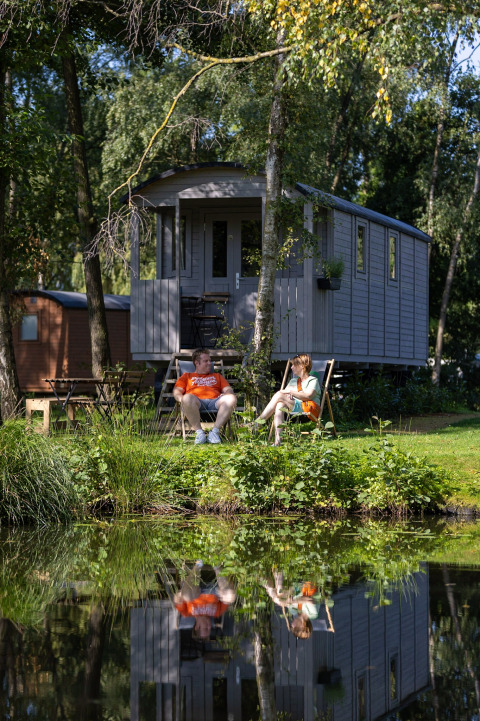 Pareja relajándose frente a una carreta gipsy con terraza en Camping Papillon, Bélgica, junto al estanque.