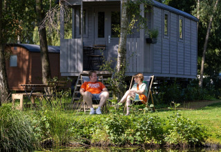 Pareja relajándose frente a una carreta gipsy con terraza en Camping Papillon, Bélgica, junto al estanque.