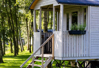Gipsy wagon cabin with terrace in a sunlit forest, surrounded by trees and grass in serene nature.