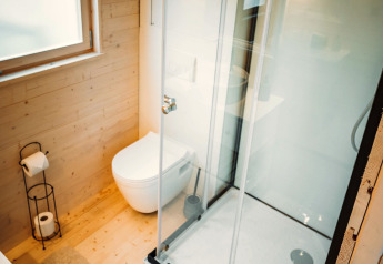 Moderne, lyst badeværelse i en træhytte med brusekabine, væghængt toilet og stort vindue med rullegardin.