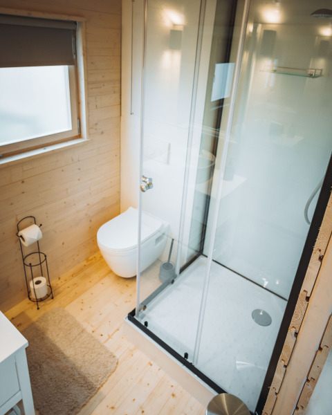 Modern badkamerinterieur in een houten hut met glazen douchecabine, hangtoilet en groot raam met rolgordijn.