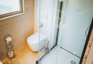 Baño contemporáneo en cabaña de madera con ducha acristalada, inodoro suspendido y ventana amplia.