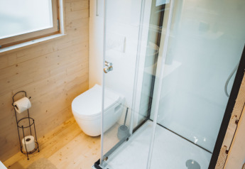 Moderne, lyst badeværelse i en træhytte med brusekabine, væghængt toilet og stort vindue med rullegardin.