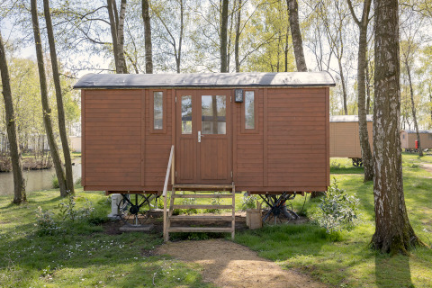 Kleine bruine pipowagen op wielen bij Camping Papillon in België, omringd door bomen en natuur.