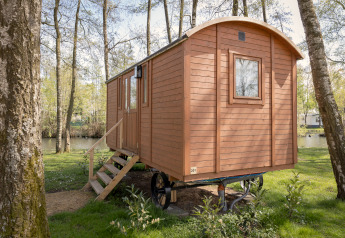 Houten Pipowagen cabin met trap tussen bomen bij een rustig meer in een groene bosrijke omgeving.