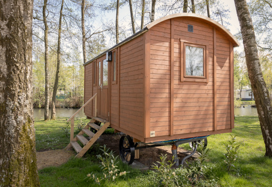 Houten Pipowagen cabin met trap tussen bomen bij een rustig meer in een groene bosrijke omgeving.