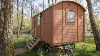 Eine kleine Holzhütte namens Pipowagen mit Treppe steht im Wald nah an einem stillen See.