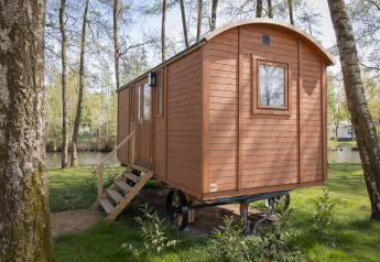 Eine kleine Holzhütte namens Pipowagen mit Treppe steht im Wald nah an einem stillen See.