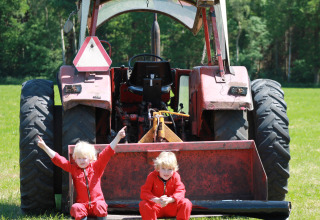 Twee kinderen in rode pakken zitten voor een rode tractor op een groene weide in zonnig Overijssel.