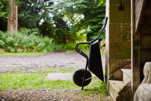 Une brouette posée contre un mur en pierre près d’un escalier, vue sur un chemin et la verdure à l’extérieur.