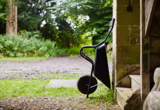 Une brouette posée contre un mur en pierre près d’un escalier, vue sur un chemin et la verdure à l’extérieur.