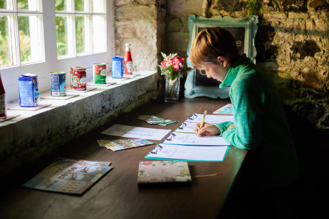 Boy writing at a rustic desk with canned goods on the windowsill at Feather Down Chesters, North West England.