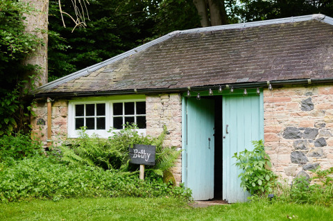 Gezellig stenen huisje met muntgroene deuren en 'Bothy'-bord te midden van weelderige begroeiing.