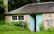 Gezellig stenen huisje met muntgroene deuren en 'Bothy'-bord te midden van weelderige begroeiing.