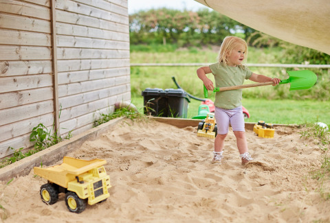 Kleines Mädchen spielt mit einer grünen Schaufel im Sandkasten bei Feather Down Heydon Grove Farm, England.