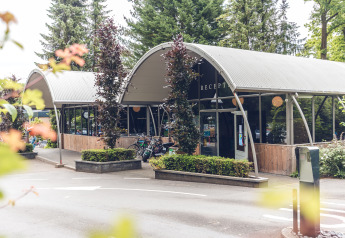 Moderne reception med buet tag ved Veluwepark de Bosgraaf, omgivet af træer og planter, cykler foran.