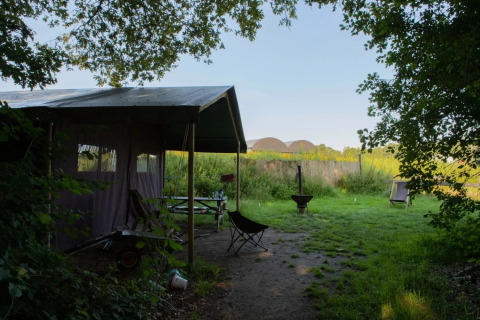 Tienda tipo safari en un campamento verde al aire libre con sillas, fogata y mesa rodeada de naturaleza.