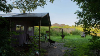 Safaritent op een groen campingterrein met stoelen, vuurplaats en tafel, omringd door natuur.