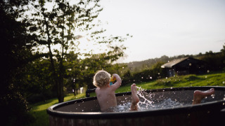 Et barn leger i en udendørs badetønde med vand, med frodige grønne marker og træer i baggrunden i solskin.