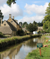 Idyllisk landsbybillede nær Malvern med traditionelle stenhuse, kanal, hundeluftere og grønt område.