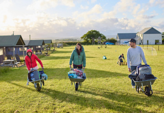 Tre personer transporterer bagage i trillebøre på Feather Down Gwel Teg Farm feriepark i det sydvestlige England.