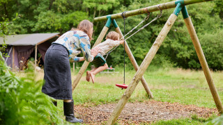 En voksen skubber et barn på en gynge i det grønne ved Feather Down Upper Shadymoor Farm i Wales, Storbritannien.