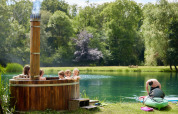 Children relax in a wooden hot tub by a lake as a man prepares a kayak at Feather Down Upper Shadymoor Farm, Wales.