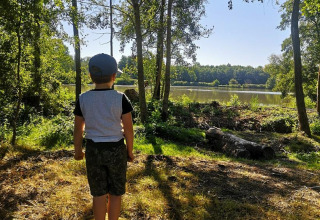 Un niño de pie descalzo sobre un tronco observa un lago en Feather Down La Ferme de Fontenille, rodeado de árboles.
